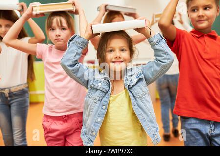 Portrait d'écolière souriante équilibrant livre sur la tête avec des camarades de classe pendant l'activité en classe Banque D'Images