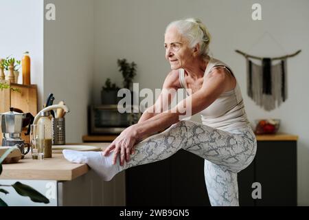 Femme âgée active faisant l'effort tout en étirant les bras sur la jambe levée sur le comptoir de cuisine et en faisant des exercices physiques le matin Banque D'Images