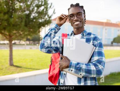 Souriant jeune étudiant noir dans des lunettes tenant un ordinateur portable à l'extérieur Banque D'Images
