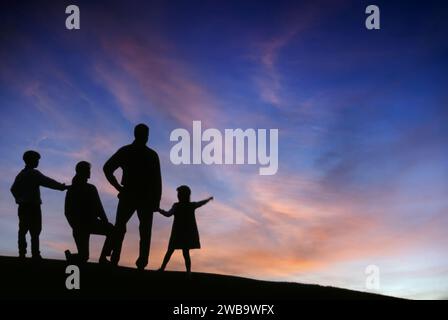 Silhouette d'une famille de quatre personnes debout sur une colline regardant de beaux nuages de coucher de soleil Banque D'Images