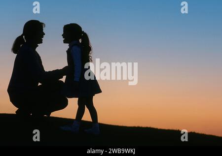 Mère et fille ayant j'ai une conversation heureuse silhouetted sur une colline et coucher de soleil Banque D'Images