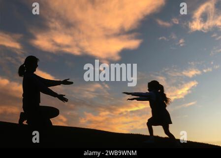 Une petite fille se profilait sur une colline et le coucher du soleil se précipitant vers ses mères attendant les bras Banque D'Images