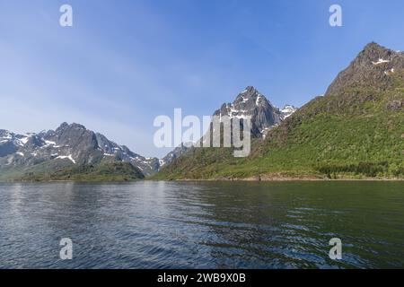 Les pics abrupts des Lofoten, avec de la neige persistante, se dressent au-dessus des eaux scintillantes d'un fjord norvégien, incarnant l'essence vibrante de l'été Banque D'Images