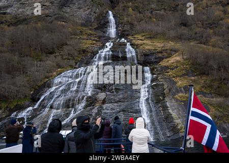 Plusieurs personnes observent depuis un bateau la cascade des sept Sœurs (de Syv Søstrene), située dans le fjord de Geiranger. Le fjord Geiranger, situé en Norvège, est un paysage naturel magnifique caractérisé par des eaux calmes entourées de montagnes escarpées. Il attire les touristes avec des activités telles que les croisières, le kayak et la randonnée. La région environnante abrite une riche variété de flore et de faune. Classée au patrimoine mondial de l'UNESCO, elle est réputée pour sa conservation et son statut d'exemple remarquable de beauté naturelle norvégienne. Banque D'Images