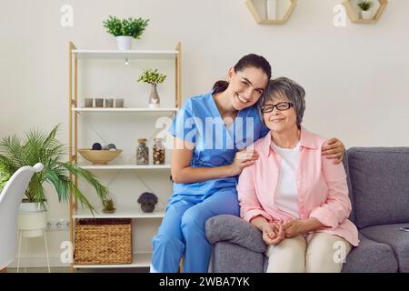 Belle, jeune, heureuse, souriante infirmière embrasse gentiment sa patiente, soignée, jolie dame aux cheveux gris. Banque D'Images