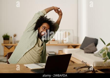 Jeune homme en vêtements décontractés levant les bras tendus au-dessus de la tête et se penchant de côté tout en étant assis devant l'ordinateur portable et profitant d'une minute de repos Banque D'Images