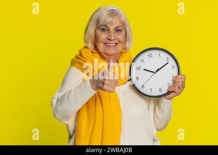 C'est ton heure, dépêche-toi ! Vieille femme âgée montrant le temps sur l'horloge de bureau mural, ok pouce vers le haut approuver pointer le doigt à la caméra, publicité, date limite. Grand-mère âgée isolée sur fond jaune Banque D'Images