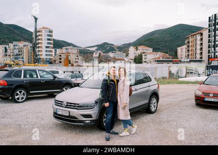 Couple souriant se tient près d'une voiture dans un parking sur fond de gratte-ciel de bâtiments au pied des montagnes Banque D'Images