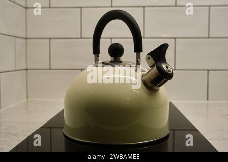 Une bouilloire sifflante en acier inoxydable de couleur crème, sur une table de cuisson à induction noire. Fond de cuisine carrelé blanc Banque D'Images