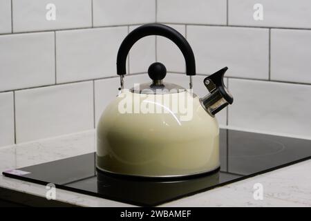 Une bouilloire sifflante en acier inoxydable de couleur crème, sur une table de cuisson à induction noire. Fond de cuisine carrelé blanc Banque D'Images