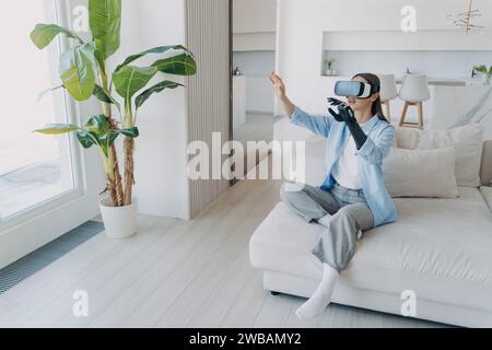 Femme émerveillée avec bras prothétique expérimentant la réalité virtuelle, faisant des gestes assis sur un canapé dans une pièce ensoleillée Banque D'Images