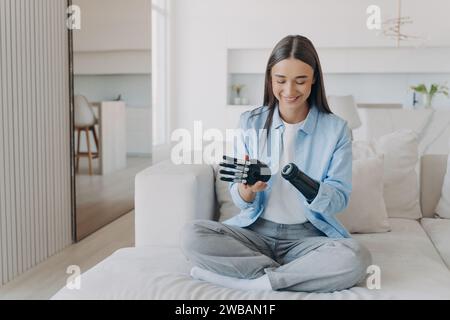 Joyeuse jeune femme avec un bras bionique assis sur un canapé, explorant la fonctionnalité de son membre prothétique dans une pièce lumineuse Banque D'Images