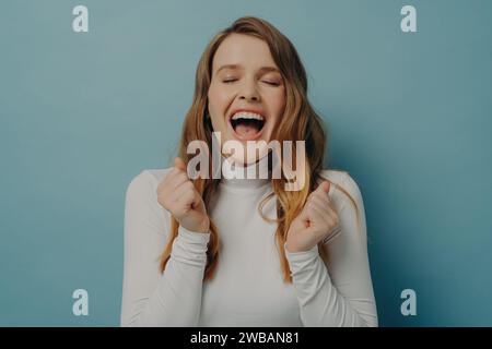 Exubérante jeune femme en col roulé blanc riant les yeux fermés, pleine de joie et de vivacité sur un fond bleu serein Banque D'Images