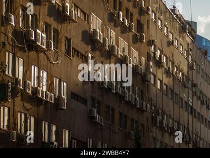 La lumière du soleil baigne une façade d'appartement parsemée d'unités de climatisation, projetant des ombres dynamiques. Banque D'Images