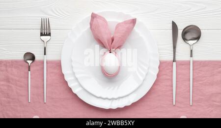 Table de vacances de Pâques avec assiette et couverts, concept d'invitation. Fond en bois blanc. Lapin fait de serviette en lin rose et oeuf. Vue de dessus. B Banque D'Images