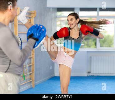 Femme athlétique dans des gants de boxe donne des coups de pied aux mitaines de punch tenues par l'entraîneur Banque D'Images