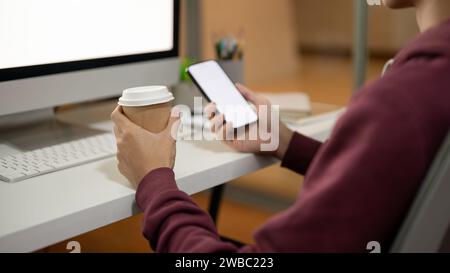 Image recadrée d'un homme de bureau utilisant son smartphone et sirotant un café à son bureau, assis devant un ordinateur. Banque D'Images