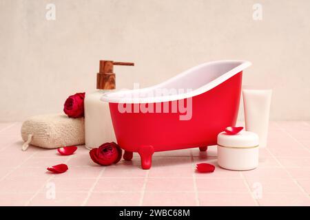 Mini baignoire avec produits cosmétiques, éponge et roses sur tuile rose près du mur blanc. Fête de la Saint-Valentin Banque D'Images