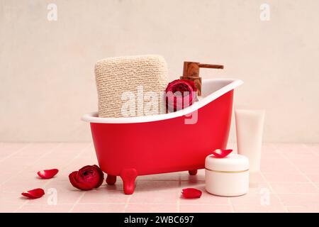 Mini baignoire avec produits cosmétiques, éponge et roses sur tuile rose près du mur blanc. Fête de la Saint-Valentin Banque D'Images