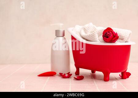 Mini baignoire avec bouteille de shampooing, serviette et roses sur tuile rose près du mur blanc. Fête de la Saint-Valentin Banque D'Images