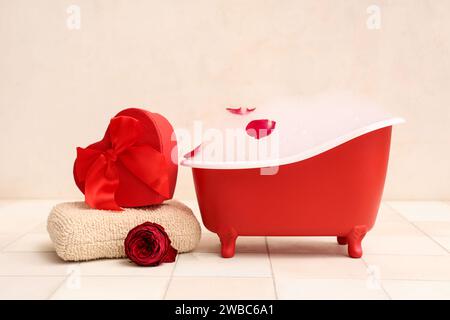 Mini baignoire avec mousse de savon, boîte-cadeau, serviette et pétales de rose sur tuile blanche près du mur. Fête de la Saint-Valentin Banque D'Images