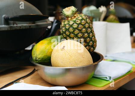 Une scène de cuisine avec un bol en acier inoxydable avec un melon mûr, papaye et ananas, prêt pour un festin de fruits tropicaux. Banque D'Images