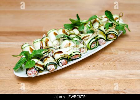 Roulés délicats à la courgette garnis de fromage infusé aux herbes et de saumon fumé, garnis de légumes verts frais, présentés sur un plateau blanc. Banque D'Images