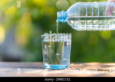 Main tenant la bouteille d'eau potable versant dans le verre sur la table en bois sur fond de nature vert flou. Eau claire et propre dans un verre Banque D'Images