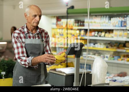 Vieux vendeur dans un magasin pesant des bananes sur des balances emballant des fruits en attente d'une vente Banque D'Images