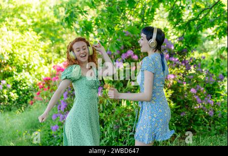 Portrait en plein air de deux femmes heureuses, amies, amies en robes et écouteurs dansant dans le parc public. Écoutez de la musique en ligne n'importe où et b Banque D'Images