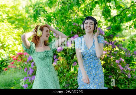 Portrait en plein air de deux femmes heureuses, amies, amies en robes et écouteurs dansant dans le parc public. Écoutez de la musique en ligne n'importe où et b Banque D'Images