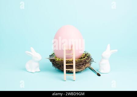 Lapin ou lapin de Pâques assis à côté d'un énorme œuf de couleur rose dans un nid d'oiseaux, pinceau et échelle, vacances de printemps Banque D'Images