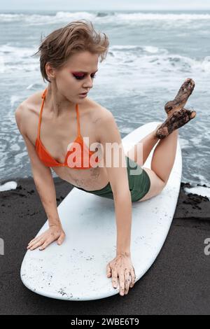 La surfeuse féminine se trouve sur le devant de la planche de surf, arquant son dos et croisant les jambes tout en se penchant sur les mains. Modèle de fitness regardant loin, faisant l'échauffement Banque D'Images