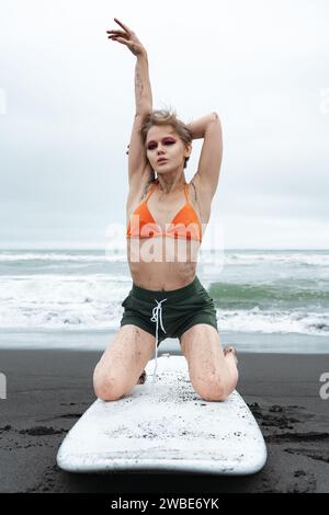 Flexibilité femme surfeuse pose sur ses genoux sur la planche de surf sur la plage de sable. surfeuse féminine a l'air incroyable en tant que mannequin de mode sportive, levant une main vers le haut Banque D'Images