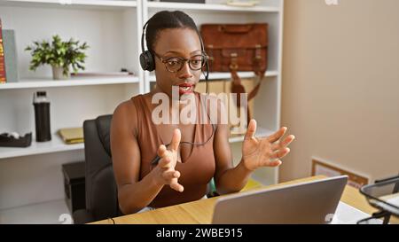 Belle femme afro-américaine patron portant des lunettes, engagé chaleureusement dans un appel d'affaires en ligne. travaillant à l'intérieur dans son bureau, elle est détendue mais s Banque D'Images