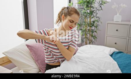 Une jeune femme grimaque dans la douleur, tenant son épaule dans une chambre lumineuse. Banque D'Images