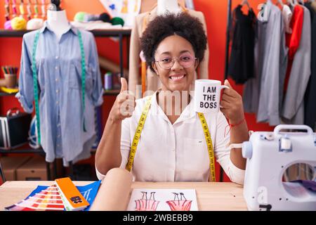 Belle femme africaine avec couturière frisée de cheveux buvant de je suis la tasse de patron souriant heureux et positif, pouce vers le haut faisant excellent et un Banque D'Images