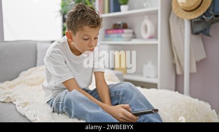 Adorable enfant blond portant une expression ennuyée, se bousculant confortablement sur le canapé à la maison, semblant sérieux tout en se relaxant et en regardant la télévision dans le salon Banque D'Images