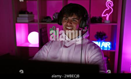 Souriant jeune homme hispanique streamer casque de sport, merveilleusement immergé dans le pouls d'un flux de jeu nocturne dans sa salle de jeu intérieure Banque D'Images