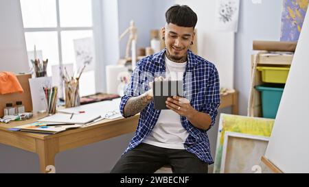 Beau jeune artiste latin en toute confiance utilisant le touchpad, souriant tout en peignant sur toile dans un studio d'art animé, incarnant la créativité et la passion. Banque D'Images