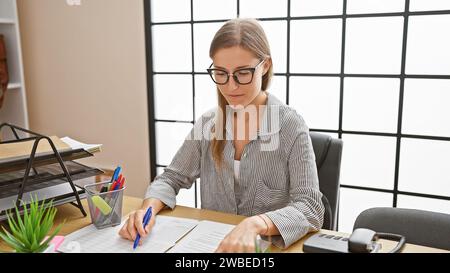 Une jeune femme caucasienne concentrée travaille avec diligence dans un bureau moderne, stylo à la main, entourée de fournitures de bureau. Banque D'Images