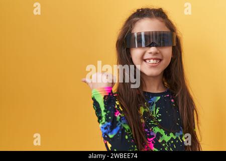 Adorable fille hispanique souriant joyeusement, regardant et pointant sur le côté avec le pouce vers le haut. porter des lunettes de réalité virtuelle. sur fond jaune isolé Banque D'Images