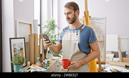 Bel homme d'âge moyen en tablier tenant le téléphone et le café dans l'atelier d'art. Banque D'Images