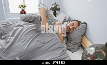 Bel homme hispanique d'âge moyen avec une barbe dormant paisiblement dans une chambre bien éclairée avec literie grise à la maison. Banque D'Images