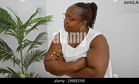 Femme afro-américaine sérieuse, un Manager réussi avec des tresses et des lunettes, assis détendu mais concentré dans la salle d'attente, les bras croisés, subtilement Banque D'Images