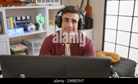 Beau jeune homme hispanique souriant en toute confiance tout en étant assis au studio de musique, écouteurs sur, absorbé dans la création de chansons, incarnant l'âme d'un Banque D'Images