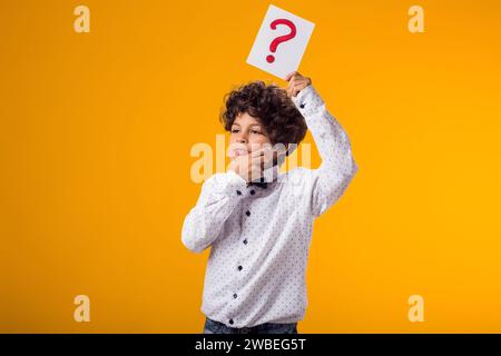 Portrait d'un enfant garçon tenant une carte de point d'interrogation sur fond jaune. Brainstorming et concept de choix Banque D'Images