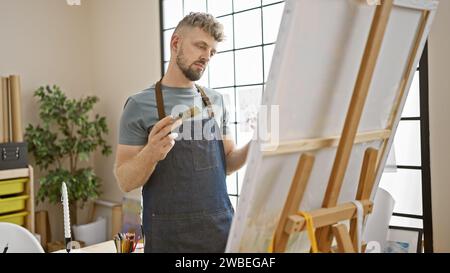 Jeune homme caucasien avec une barbe et les yeux bleus, portant un tablier en denim, peignant avec soin sur un chevalet dans un studio lumineux Banque D'Images