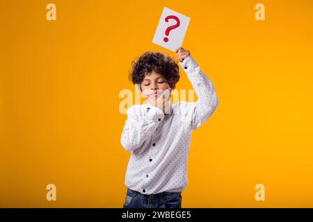 Portrait d'un enfant garçon tenant une carte de point d'interrogation sur fond jaune. Brainstorming et concept de choix Banque D'Images