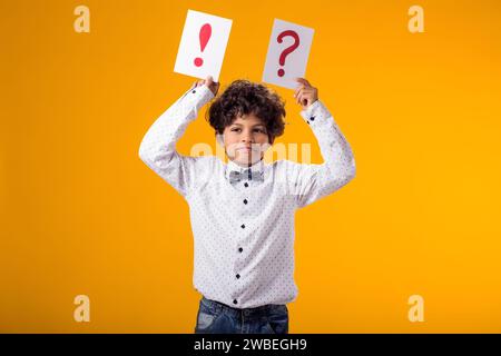 Portrait d'un enfant garçon tenant des cartes de point d'interrogation et de point d'exclamation sur fond jaune. Brainstorming et concept de choix Banque D'Images